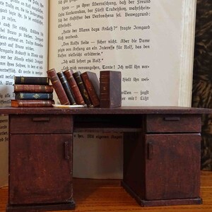 May include: A miniature wooden desk with two drawers and a door. The desk is brown and has a worn, rustic look. There are several books stacked on the desk, and a book is open on the left side of the desk.