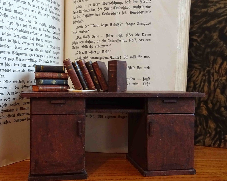May include: A miniature wooden desk with two drawers and a door. The desk is brown and has a worn, rustic look. There are several books stacked on the desk, and a book is open on the left side of the desk.