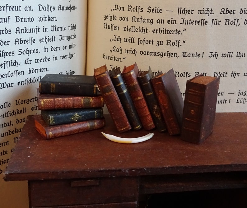 May include: A collection of miniature antique books with brown leather covers and gold accents. The books are arranged on a wooden surface with a bone-colored object in the foreground.