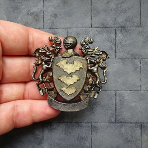 Puede incluir: Un escudo de armas de metal dorado y negro con un casco de caballero y tres murciélagos en el escudo.