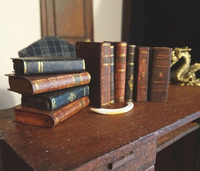 May include: A collection of miniature antique-style books with gold accents, stacked on a wooden table. The books are arranged in a row, with the spines facing the viewer. The books are all different sizes and have different titles.
