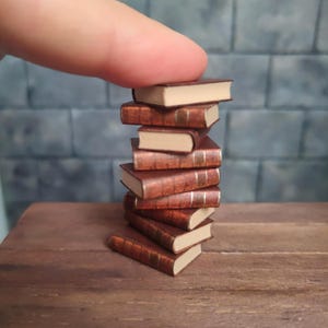 May include: A stack of miniature books with brown covers and cream-coloured pages. The books are arranged in a slightly tilted tower, with gold accents on the spines. The books are on a wooden surface.