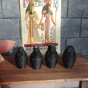 Puede incluir: Cuatro pequeños frascos canópicos en miniatura de color negro con diseños jeroglíficos. Los frascos están sobre una mesa de madera delante de un cartel con temática egipcia.