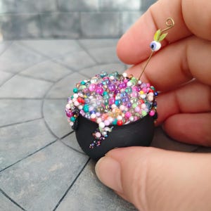 May include: Miniature black cauldron overflowing with colorful beads. A gold wire with a green leaf and a blue and white eyeball is inserted into the cauldron. The cauldron is held in a hand.