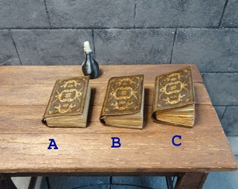 May include: Three miniature books labeled A, B, and C, each featuring a dark brown cover with intricate gold filigree patterns and aged, off-white pages. A small, dark gray bottle with a white stopper sits behind the books.