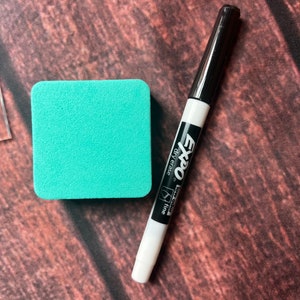 May include: A white dry-erase marker with black lettering that reads "Expo" "low odor" "fine" "dry erase". The marker is next to a turquoise square dry-erase board.