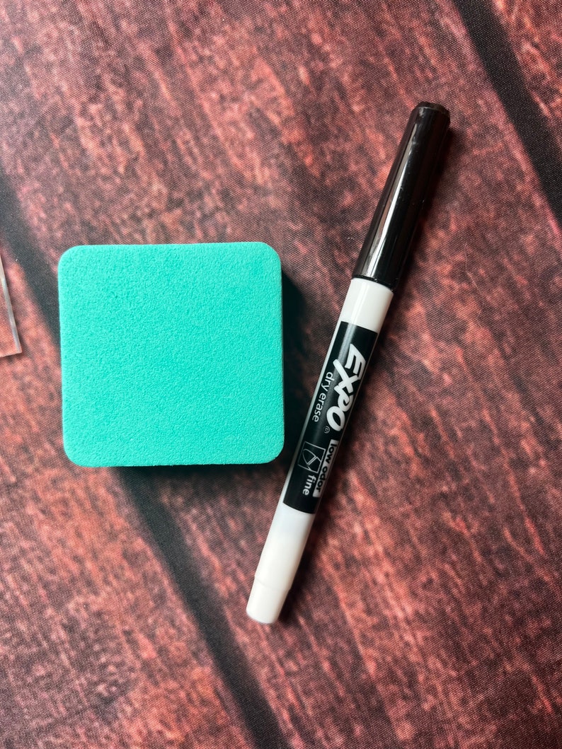 May include: A white dry-erase marker with black lettering that reads "Expo" "low odor" "fine" "dry erase". The marker is next to a turquoise square dry-erase board.