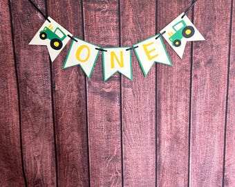 May include: A banner with the word "ONE" in yellow letters, with a green tractor on each end. The banner is white with a green border and is strung on a black string. The background is a brown wooden surface.