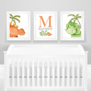 May include: Three framed prints with a dinosaur theme. The first print features a green triceratops with a palm tree. The second print features the letter M in orange with the name Matthew below it and a row of smaller dinosaurs. The third print features an orange stegosaurus with a palm tree.