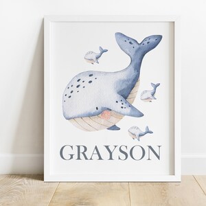 May include: A watercolor illustration of a blue whale with smaller whales swimming around it. The whale has a light blue body with dark blue spots and a light brown belly. The name "GRAYSON" is written in gray text below the whale.
