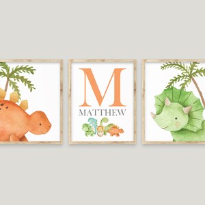 May include: Three framed prints with a dinosaur theme. The left print features a brown dinosaur with green palm trees. The middle print features the letter M in orange with the name Matthew below. The right print features a green dinosaur with green palm trees.