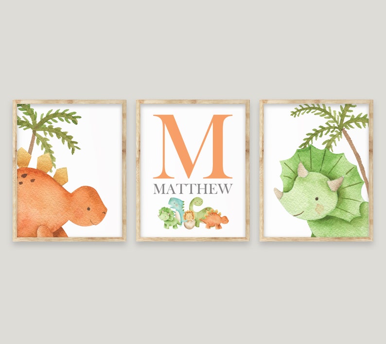May include: Three framed prints with a dinosaur theme. The left print features a brown dinosaur with green palm trees. The middle print features the letter M in orange with the name Matthew below. The right print features a green dinosaur with green palm trees.