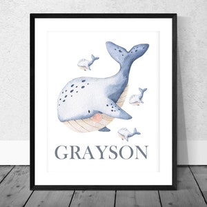 May include: A watercolor print of a blue whale with white spots and a light pink belly. The whale is surrounded by four smaller whales. The print has the name "GRAYSON" written in gray text at the bottom.