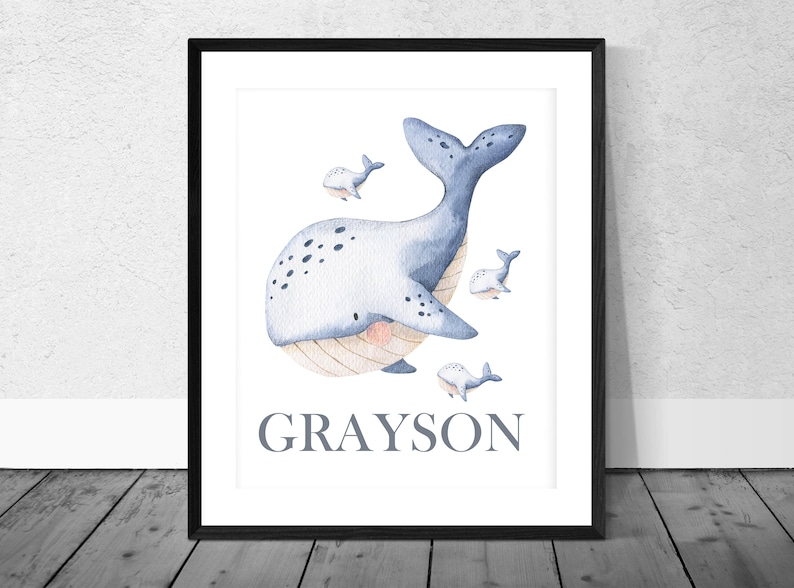 May include: A watercolor print of a blue whale with white spots and a light pink belly. The whale is surrounded by four smaller whales. The print has the name "GRAYSON" written in gray text at the bottom.