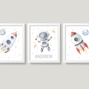 May include: Three framed watercolor prints with a space theme. The prints feature a rocket ship, an astronaut, and a rocket ship with a planet in the background. The astronaut print has the name "ANDREW" written below the astronaut.