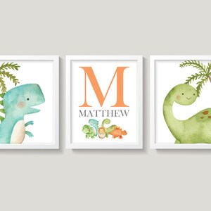 May include: Three framed prints featuring watercolor illustrations of dinosaurs. The center print has a large orange letter "M" and the name "MATTHEW" in gray text. The left print shows a blue dinosaur with a palm tree. The right print shows a green dinosaur with a palm tree.