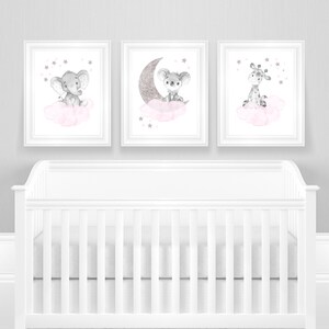 Op de afbeelding: Drie ingelijste prints met aquarel illustraties van een grijze olifant, koala en giraffe die op roze wolken zitten met zilveren sterren. De prints hangen aan een grijze muur boven een wit wiegje.