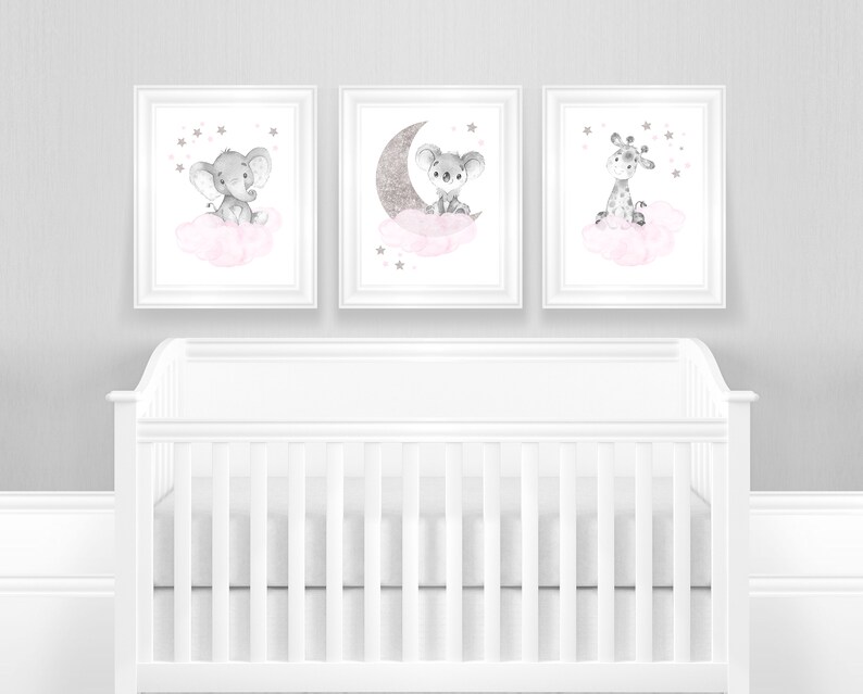 Op de afbeelding: Drie ingelijste prints met aquarel illustraties van een grijze olifant, koala en giraffe die op roze wolken zitten met zilveren sterren. De prints hangen aan een grijze muur boven een wit wiegje.