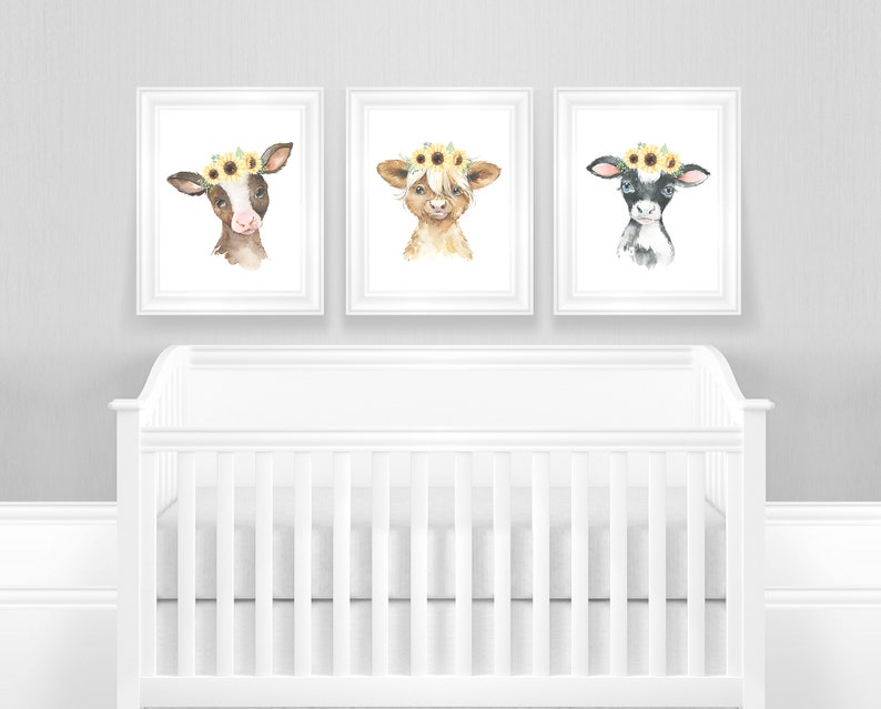 May include: Three watercolor paintings of calves wearing sunflower crowns. The calves are brown and white, brown, and black and white. The paintings are framed in white frames and are hanging on a gray wall above a white crib.