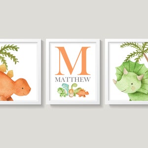 May include: Three framed prints featuring watercolor illustrations of dinosaurs. The first print shows an orange dinosaur with a palm tree behind it. The second print shows the letter M in orange with the name Matthew below it and a row of smaller dinosaurs. The third print shows a green dinosaur with a palm tree behind it.