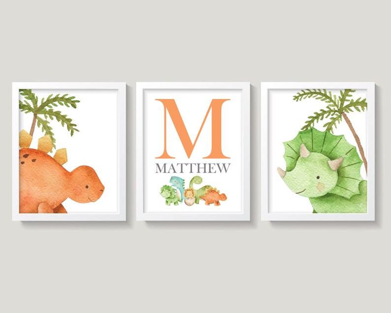 May include: Three framed prints featuring watercolor illustrations of dinosaurs. The first print shows an orange dinosaur with a palm tree behind it. The second print shows the letter M in orange with the name Matthew below it and a row of smaller dinosaurs. The third print shows a green dinosaur with a palm tree behind it.