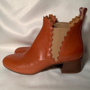 May include: A rust-colored leather ankle boot featuring a rounded toe, a low block heel, and a pull tab at the back. The boot is designed with a distinctive scalloped edge along the collar and the side elastic gore panel, which is beige.