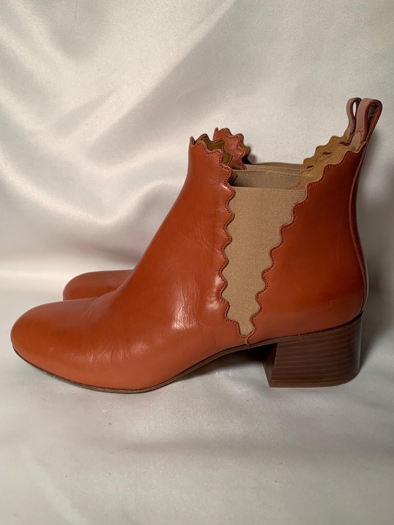 May include: A rust-colored leather ankle boot featuring a rounded toe, a low block heel, and a pull tab at the back. The boot is designed with a distinctive scalloped edge along the collar and the side elastic gore panel, which is beige.