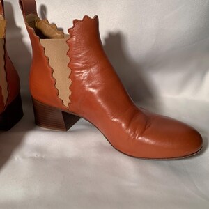 May include: A pair of cognac-colored leather ankle boots featuring a rounded toe, a stacked block heel, and a pull tab at the back. The side panels are made of beige elastic fabric with a distinct scalloped edge detail along the boot opening.