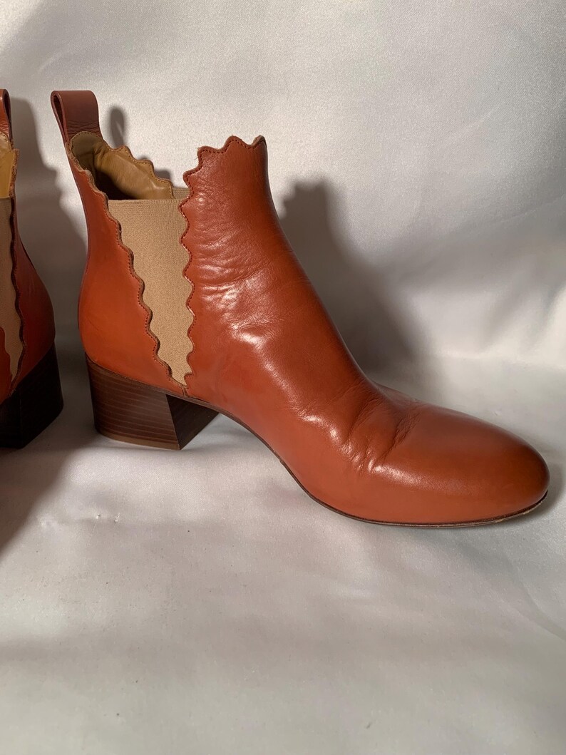 May include: A pair of cognac-colored leather ankle boots featuring a rounded toe, a stacked block heel, and a pull tab at the back. The side panels are made of beige elastic fabric with a distinct scalloped edge detail along the boot opening.