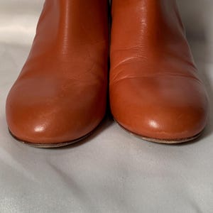May include: A pair of ankle boots crafted from smooth, cognac-colored leather. The boots feature a rounded toe design, a clean silhouette without visible laces or zippers, and a low-profile sole, showcasing a polished and uniform finish throughout.