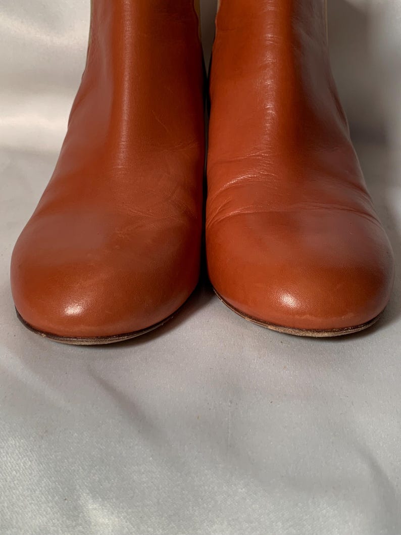 May include: A pair of ankle boots crafted from smooth, cognac-colored leather. The boots feature a rounded toe design, a clean silhouette without visible laces or zippers, and a low-profile sole, showcasing a polished and uniform finish throughout.
