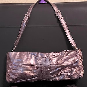May include: A metallic silver clutch with a matching shoulder strap. The bag has a textured, gathered design and a central flap with a buckle. The strap is adjustable with silver buckles. A purple object is holding the strap.