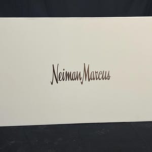 Puede incluir: Una caja rectangular blanca con las palabras "Neiman Marcus" impresas en una escritura marrón oscuro. La caja está sobre una superficie negra.