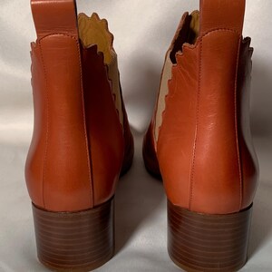 May include: A pair of rust-colored leather ankle boots featuring a scalloped edge along the side panels, tan elastic gussets, a vertical pull tab at the heel, and a stacked brown block heel. The boots are shown from the back view.