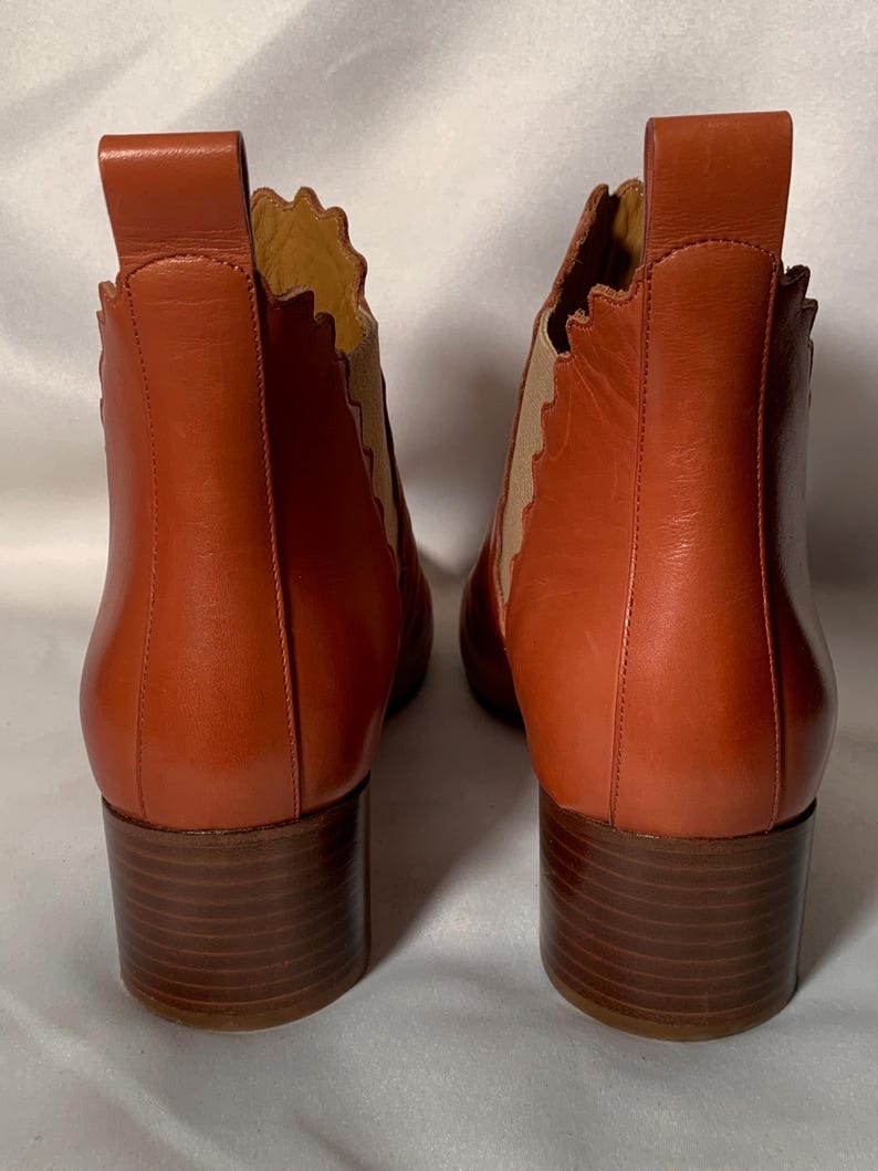 May include: A pair of rust-colored leather ankle boots featuring a scalloped edge along the side panels, tan elastic gussets, a vertical pull tab at the heel, and a stacked brown block heel. The boots are shown from the back view.