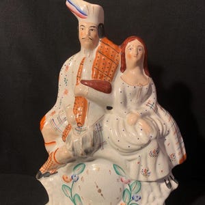 Könnte beinhalten: Eine Keramik-Kaminuhr mit einer handbemalten Figur eines Paares in traditioneller schottischer Kleidung. Das Zifferblatt ist in den Sockel integriert und mit grünen Blättern und kleinen Blumen verziert. Die Figuren tragen weiße Kleidung mit orangefarbenen und blauen Karomustern.