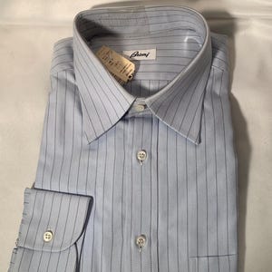May include: Light blue dress shirt with vertical navy pinstripes. The shirt has a classic collar, buttoned cuffs, and a chest pocket. The brand name "Brioni" is visible on the label.