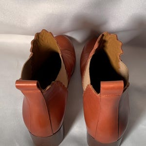 May include: A pair of cognac-colored leather ankle boots featuring a scalloped topline, a vertical pull tab at the heel, and a tan elastic side panel. The boots have a stacked block heel and a smooth finish, viewed from a top-down perspective.