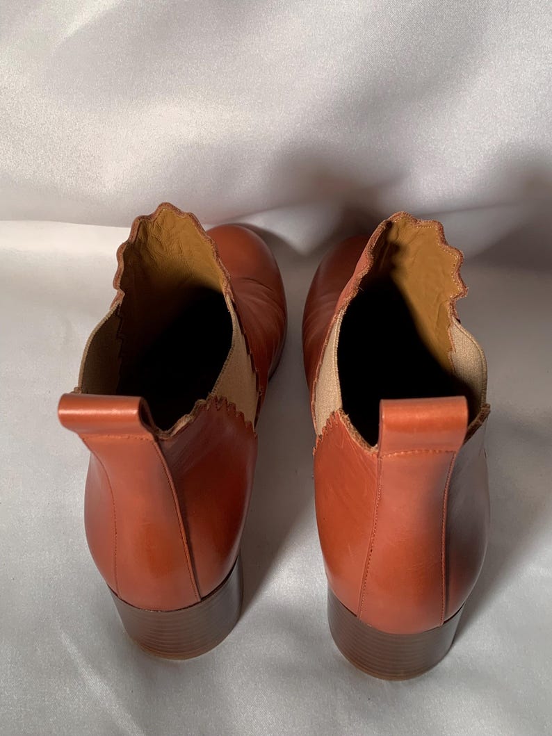 May include: A pair of cognac-colored leather ankle boots featuring a scalloped topline, a vertical pull tab at the heel, and a tan elastic side panel. The boots have a stacked block heel and a smooth finish, viewed from a top-down perspective.