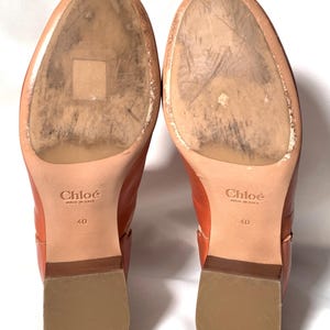 May include: A pair of tan leather boots with worn soles, featuring the Chlo&eacute; logo and the text Made in Italy and 40 stamped on the bottom. The boots have a low block heel with a light brown finish and a smooth, rounded toe area on the sole.