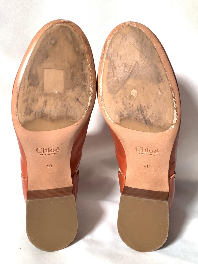 May include: A pair of tan leather boots with worn soles, featuring the Chlo&eacute; logo and the text Made in Italy and 40 stamped on the bottom. The boots have a low block heel with a light brown finish and a smooth, rounded toe area on the sole.
