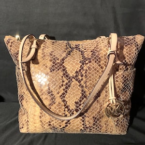 Puede incluir: Bolso tote beige y marrón con estampado de piel de serpiente, con cremallera y asas doradas. El bolso tiene un charm con el logotipo MK dorado y una correa beige claro. Hay un bolsillo lateral.
