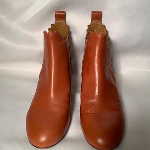 May include: A pair of cognac-colored leather ankle boots featuring a rounded toe, a pull tab at the heel, and a distinctive scalloped edge along the top opening. The boots include elastic side panels in a lighter tan shade for a flexible fit.