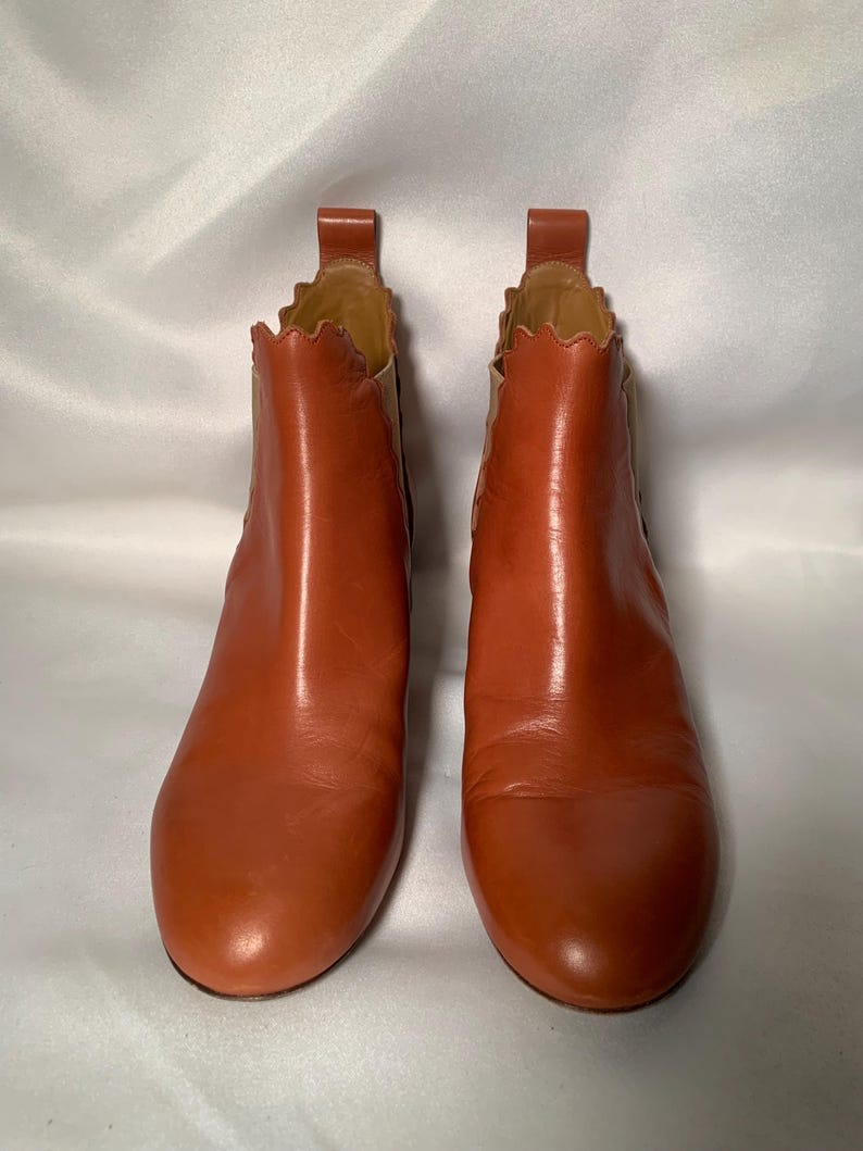 May include: A pair of cognac-colored leather ankle boots featuring a rounded toe, a pull tab at the heel, and a distinctive scalloped edge along the top opening. The boots include elastic side panels in a lighter tan shade for a flexible fit.