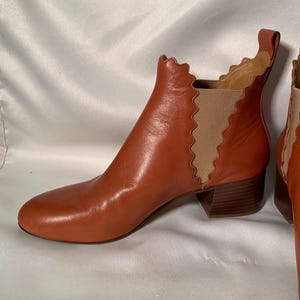 May include: A cognac-colored leather ankle boot featuring a rounded toe, a low block heel, and a distinctive scalloped edge along the side panel. The boot includes a tan elastic gore insert for flexibility and a pull tab at the top rear collar.