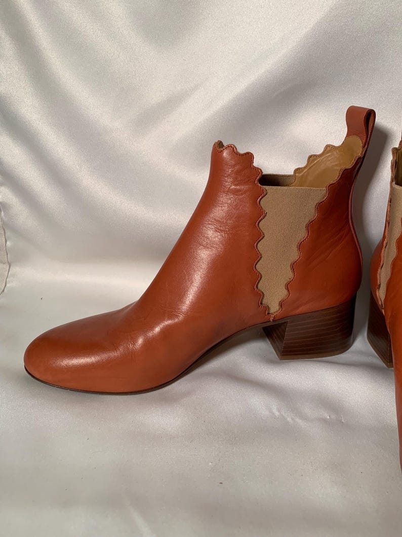 May include: A cognac-colored leather ankle boot featuring a rounded toe, a low block heel, and a distinctive scalloped edge along the side panel. The boot includes a tan elastic gore insert for flexibility and a pull tab at the top rear collar.