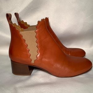 May include: A pair of cognac-colored leather ankle boots featuring a rounded toe, a block heel, and a pull tab at the back. The side panels are made of tan elastic fabric with a distinct scalloped edge detail along the leather trim.