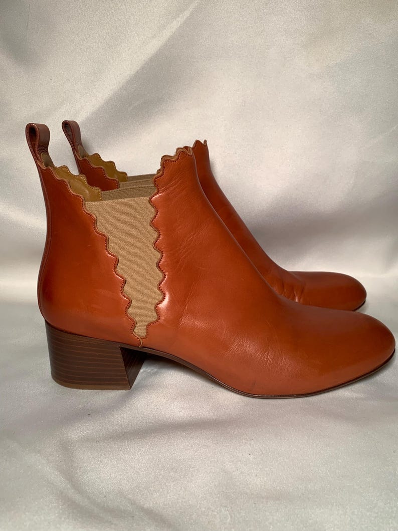 May include: A pair of cognac-colored leather ankle boots featuring a rounded toe, a block heel, and a pull tab at the back. The side panels are made of tan elastic fabric with a distinct scalloped edge detail along the leather trim.