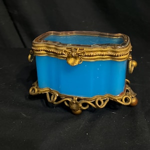 Puede incluir: Un joyero rectangular antiguo con un cuerpo azul vibrante y adornos de metal dorado ornamentados. La caja presenta una tapa de cristal transparente y detalles florales decorativos en las patas y el cierre.