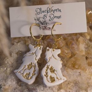 Puede incluir: Pendientes blancos y dorados con diseño de lobo y detalles florales. Los pendientes cuelgan de aros dorados. El producto se exhibe en una tarjeta blanca con el texto "Blackthorn & Bone" y una ilustración floral.